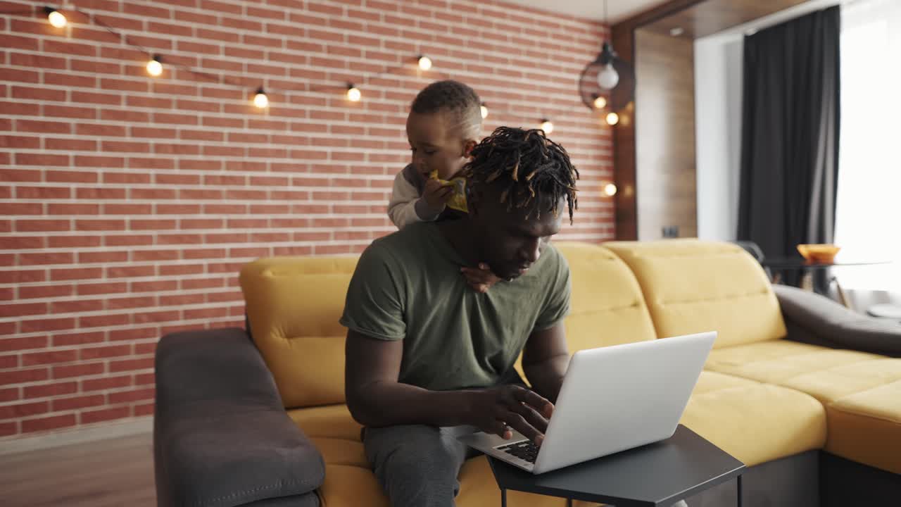 Busy young african father doing freelance work on laptop while his son disturbing him from the back