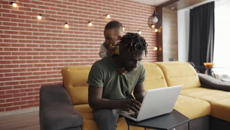 Busy-young-african-father-doing-freelance-work-on-laptop-while-his-son-disturbing-him-from-the-back