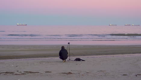 el técnico de sonido agazapado está grabando ambientes en la playa al atardecer, el cielo rosa y las olas del mar con el horizonte al fondo de la secuencia