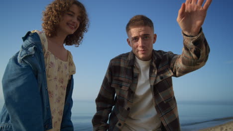 Happy-couple-waving-hands-saying-hello-on-camera-standing-sandy-beach.