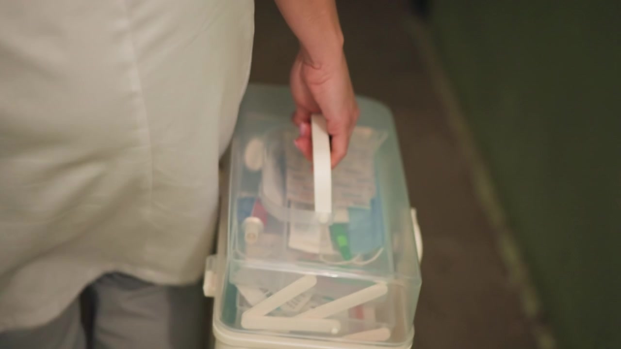 Infermiera che trasporta un kit medico lungo un corridoio di notte, operatrice sanitaria in uniforme bianca afferra una scatola di forniture trasparente piena di bende e strumenti, corridoio domestico fiocamente illuminato, movimento silenzioso e concentrato