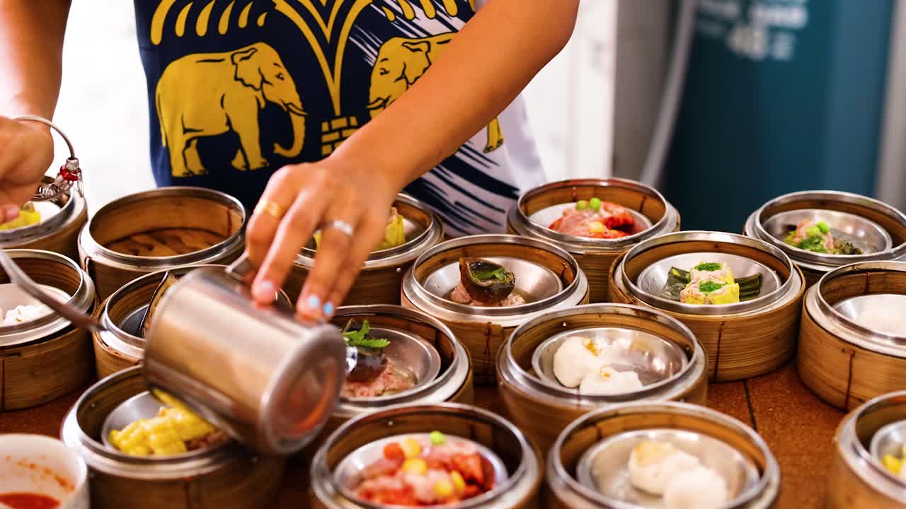 A vendor arranges and pours liquid over dim sum baskets in a vibrant Phuket market. Bright lighting highlights the colorful ingredients
