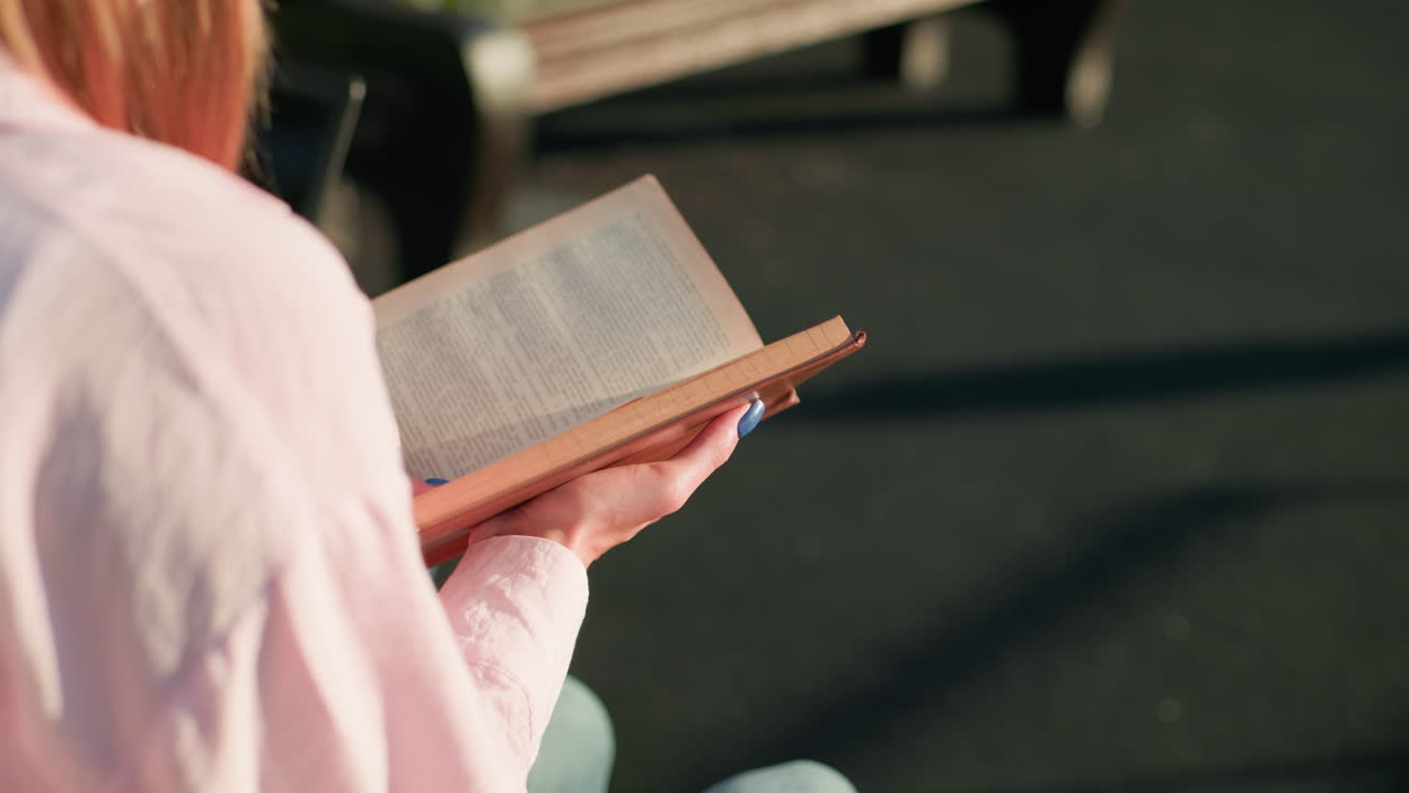 핑크색 을 입은 여자가 밖에서 앉아서 책을 들고 있는 모습, 눈에 보이는 그림자와 배경에 흐릿한 벤치