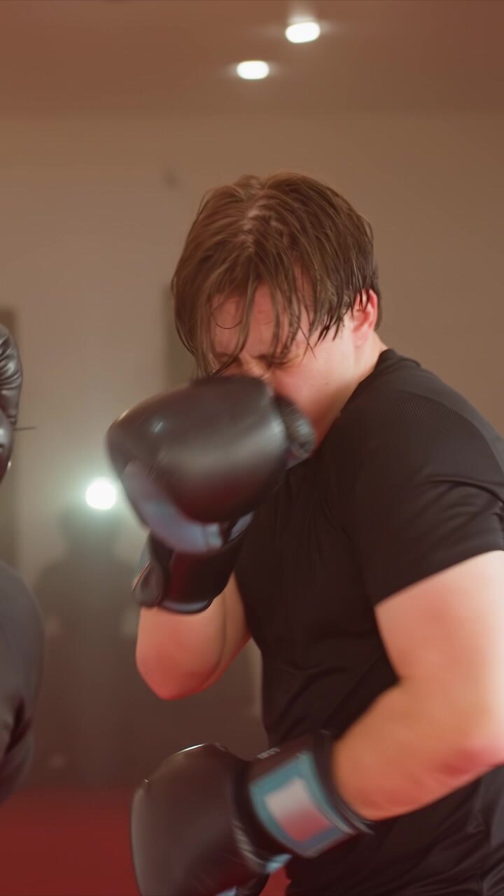 Primo piano di un giovane uomo che pratica la boxe indossando guanti neri. I guanti si muovono dinamicamente mentre l'uomo sferra pugni. Pareti beige e pavimento rosso visibili durante tutta la sessione di allenamento.