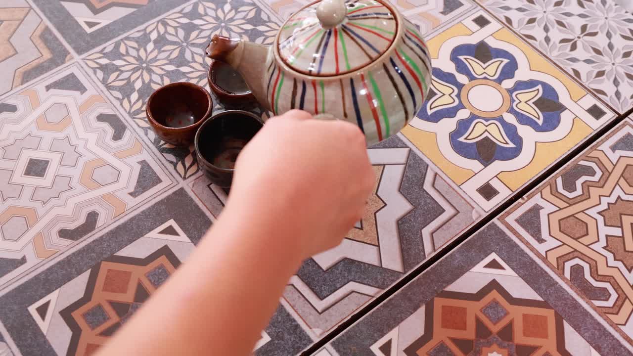 A person pours tea from a colorful teapot into small cups on a patterned table in Phuket, Thailand
