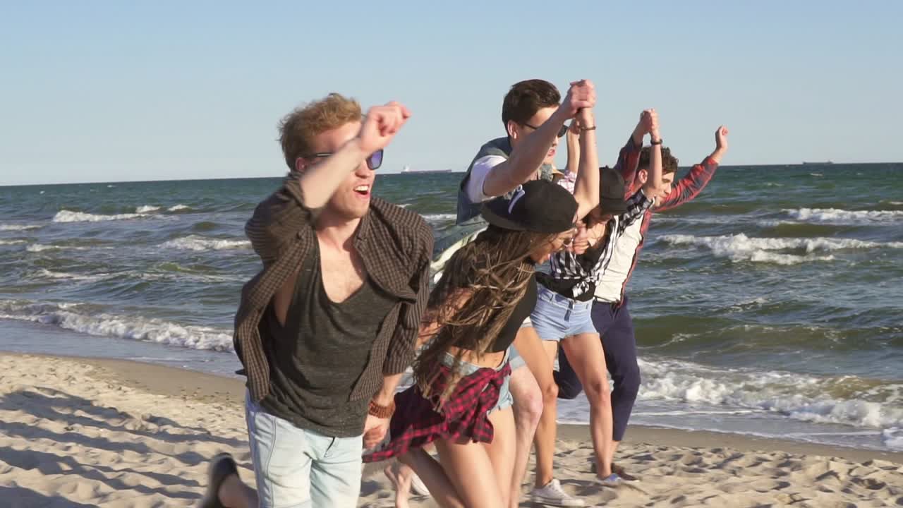 Group of young hipster friends running together holding hands on a beach at the water's edge. Slowmotion shot