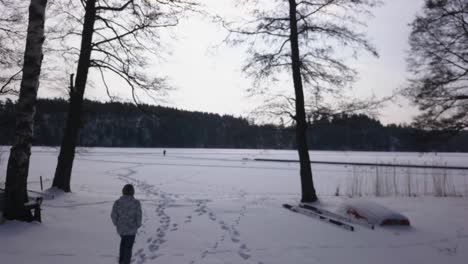 Follow-aerial-shot-of-blonde-woman-walking-towards-frozen-lake-admiring-the-snow-covered-wide-open-vista