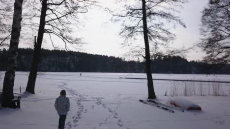 follow aerial shot of blonde woman walking towards frozen lake admiring the snow covered wide open vista