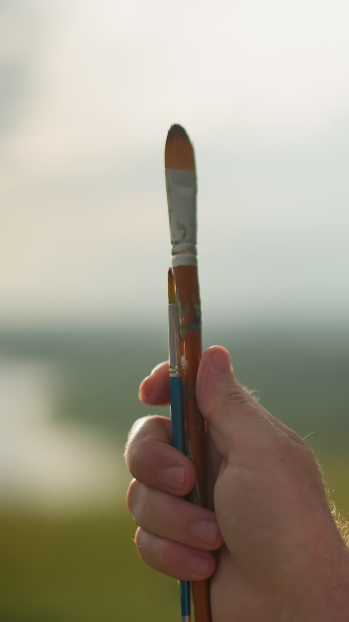 A close-up of a person's hand holding two paintbrushes one small and one very small while turning them slowly. The face is partially visible against a blurred, sunny background