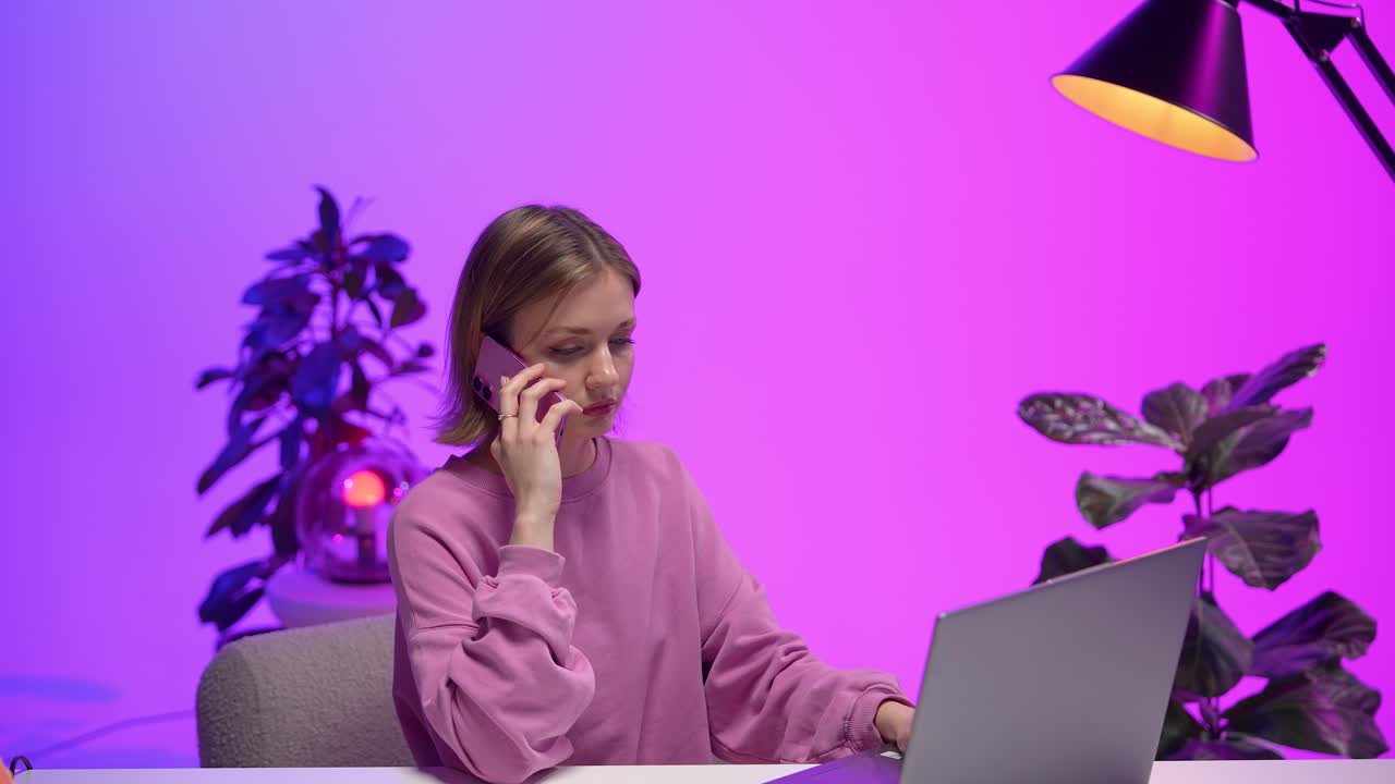 Focused young woman in a pink sweatshirt sits at a modern desk using her smartphone, laptop open, surrounded by indoor plants and vibrant magenta and violet lighting in a stylish home office