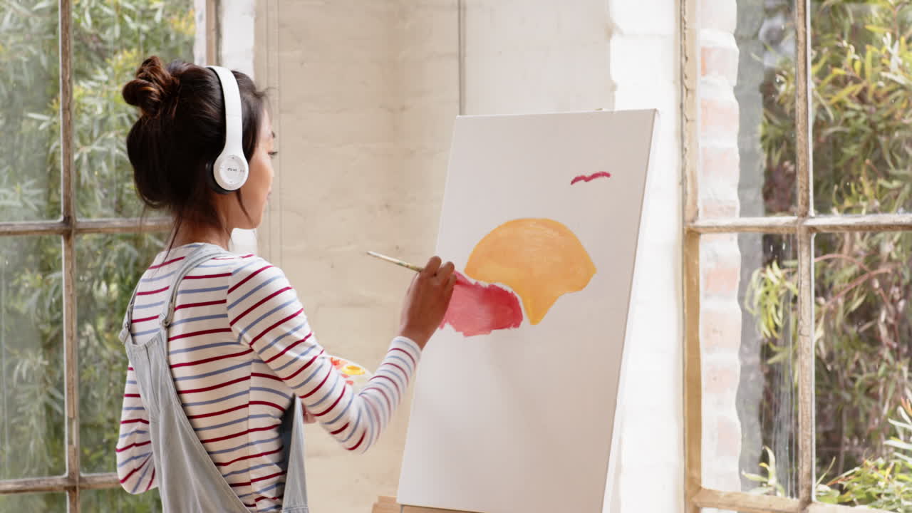 Asian young woman painting on canvas while listening to music at home studio