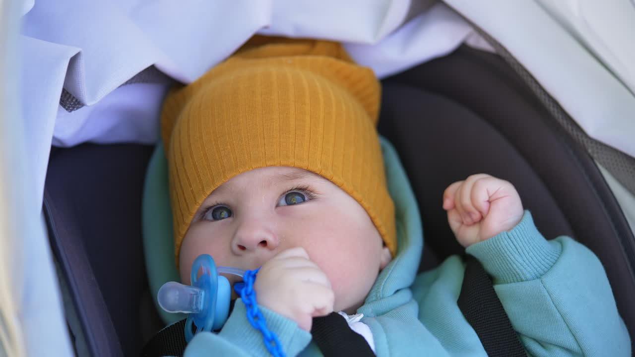 Adorable grey-eyed baby in yellow cap and blue blazer sucking thumb. Sweet kid looks up lying in the stroller outdoors. Close up.