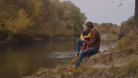 happy-father-is-communicating-with-his-little-son-at-nature-man-and-boy-are-sitting-on-coast-of-forest-river-together-at-weekend-happiness-and-love-of-dad