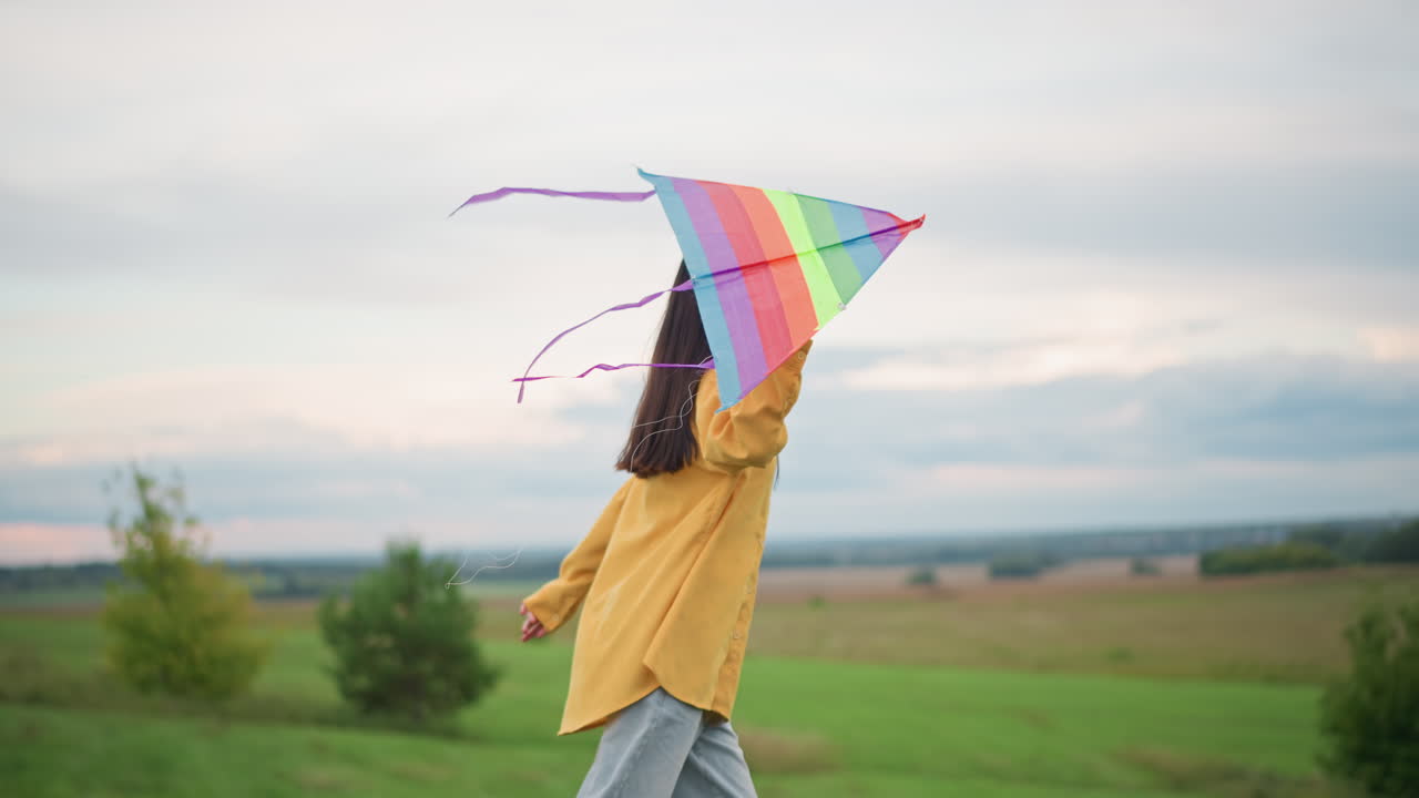 Menina voando com uma pipa colorida, criança correndo por uma paisagem verdejante com uma pipa vibrante, mulher de casaco amarelo perseguindo uma pipa de arco-íris vívido por campos gramados sob um céu nublado