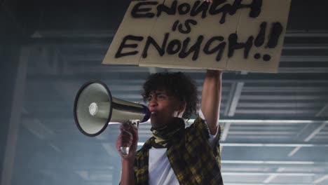 hombre afroamericano con máscara facial hablando en megáfono sosteniendo un cartel de protesta en un estacionamiento vacío