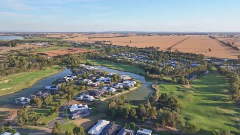 Vista-Aérea-De-Casas-Con-Obstáculos-De-Agua-Y-Calles-Del-Campo-De-Golf-Con-Campos-De-Trigo-Más-Allá-Cerca-De-Yarrawonga