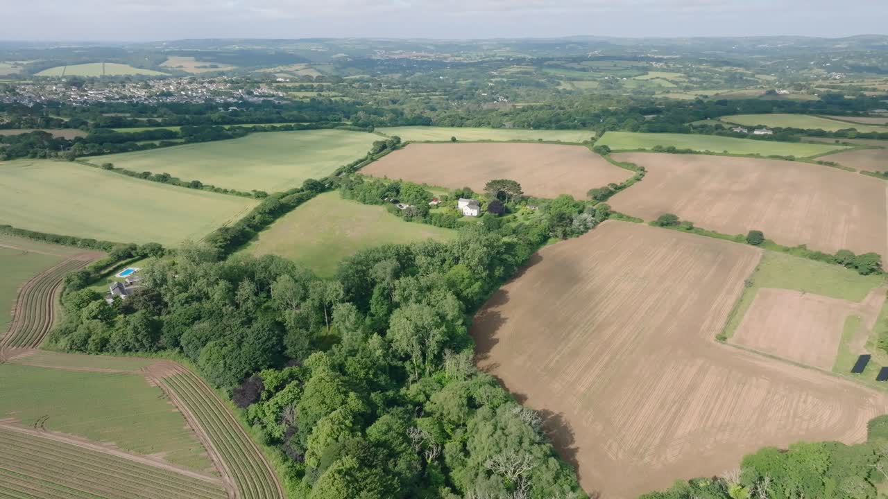 Farmhouses nestled in vibrant green woodland surrounded by farmed fields lined with hedgerows. Summer, Cornwall, UK.