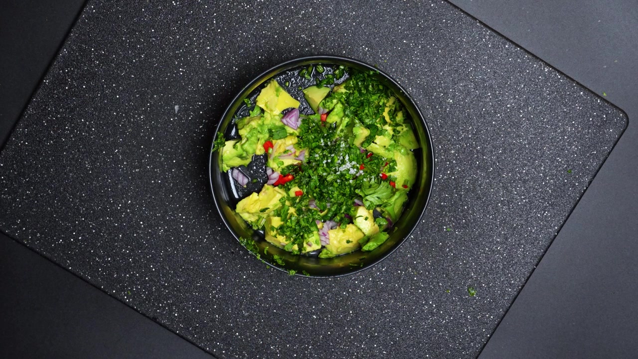 Vista dall'alto degli ingredienti del guacamole in una ciotola nera. Mani che tengono una ciotola contenente avocado a cubetti, cipolla rossa, coriandolo e peperoncino su un tagliere scuro