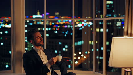 man drinking coffee and looking out a window at a city at night