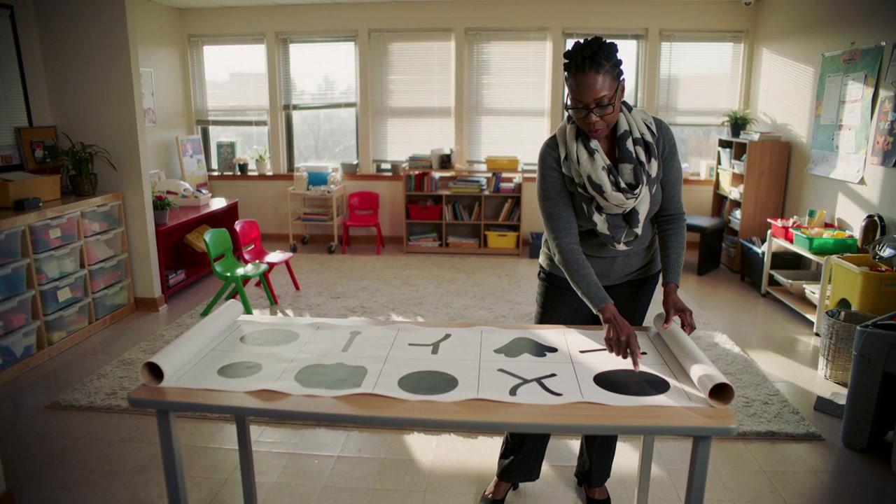Walking teacher in scarf unrolling paper with shapes on classroom table for class, pointing