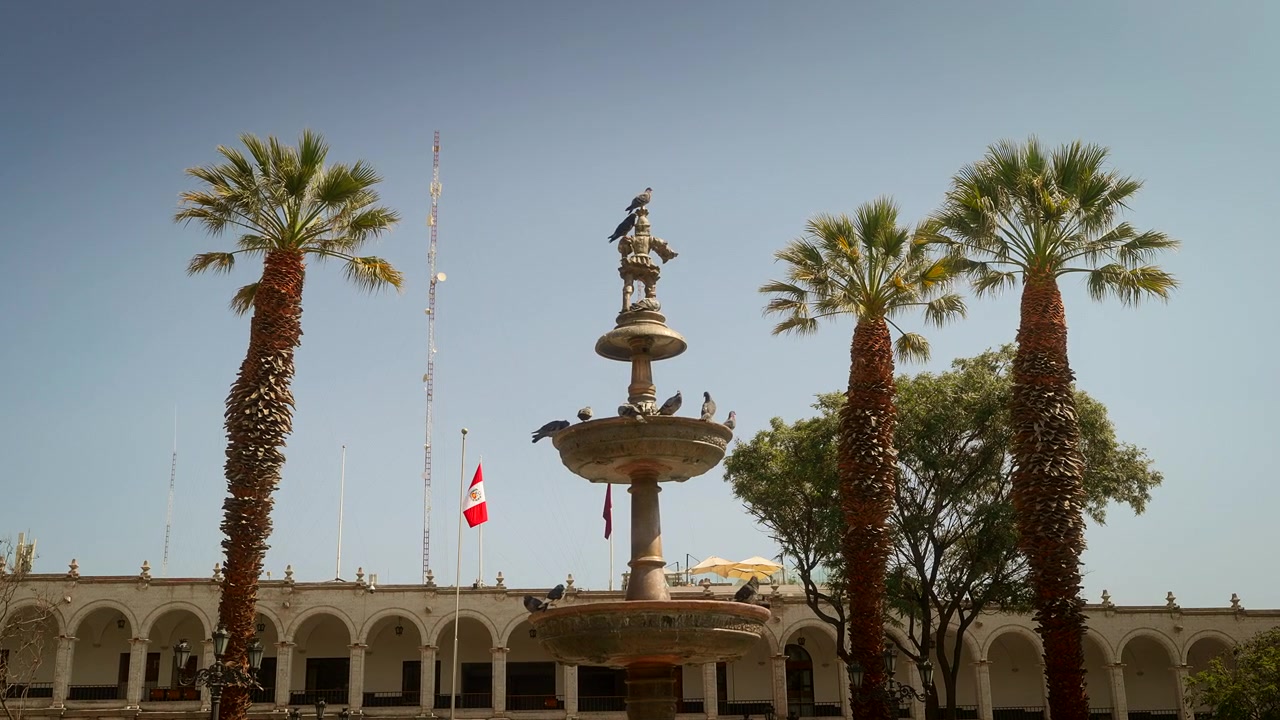 Fontana e palme in Plaza de Armas sotto cieli sereni nel centro di Arequipa, Perù, bandiere sventolano e uccelli si radunano intorno alla statua del trombettiere El Tuturutu.
