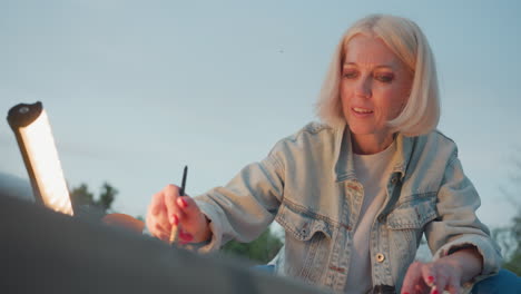 modern blonde woman drawing detailed sketch under yellow lamp glow on wooden surface outdoors at dusk, close up focused expression capturing brushstroke flow and atmosphere in soft evening light