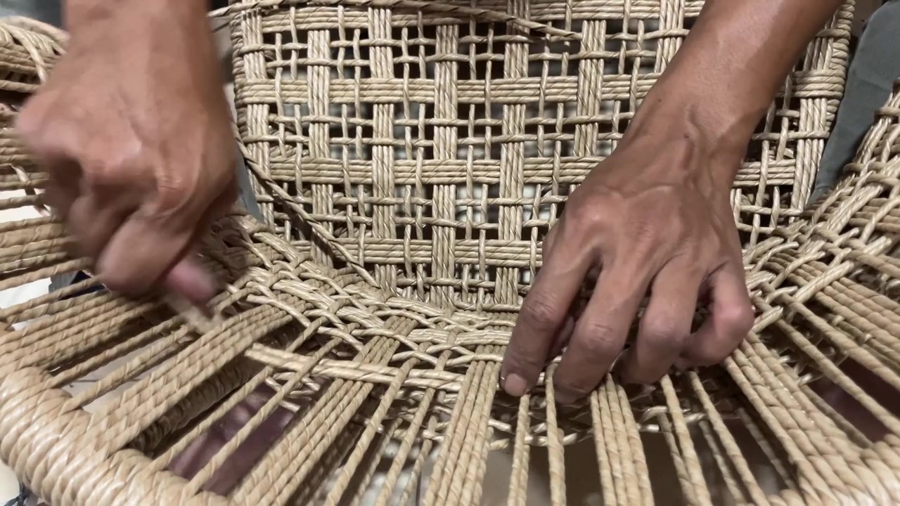 Nærbillede i slow motion af håndværkerhænder, der fletter naturfibersnor for at skabe et traditionelt stolesæde. Detaljeret visning af håndværket og processen med at fremstille håndlavede møbler.