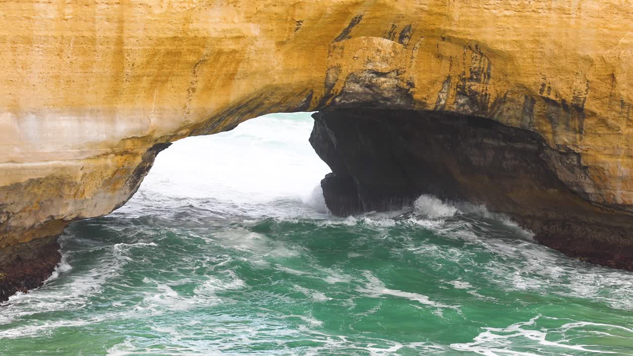 Dynamic ocean waves surge through the natural arch of London Bridge, Port Campbell, under soft natural lighting