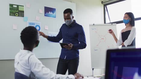 african american man and woman wearing face masks giving presentation to their colleagues in meeting