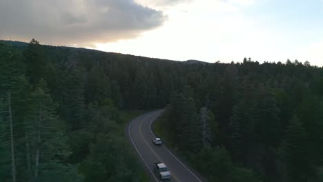 drone shot of white car moving on countryside road surrounded by thick evergreen forest in dusk