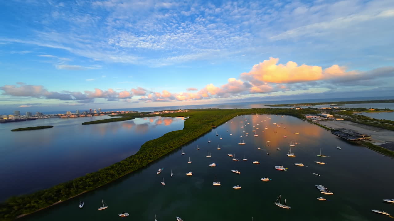 Bay with multiple yachts, green islands in the vast waterscape. Urban skyline at backdrop. Sky with colorful clouds low angle view. FPV drone footage.