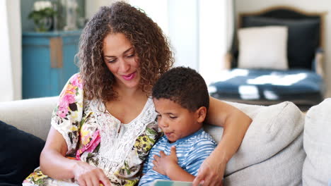 Toma-En-Cámara-Lenta-De-Una-Madre-Sentada-En-Un-Sofá-En-Casa-Leyendo-Un-Libro-Con-Su-Hijo