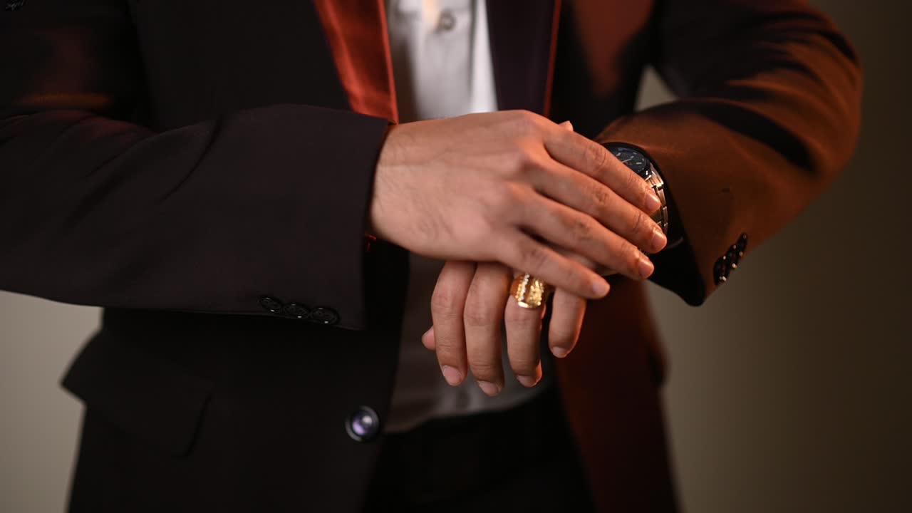 Un hombre con una chaqueta de esmoquin formal, camisa blanca y un anillo de oro revisa su reloj de pulsera. La iluminación cinematográfica resalta las texturas de la tela y las joyas.