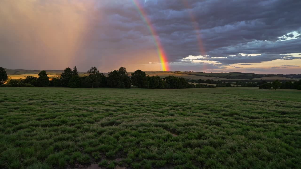 The video captures a serene landscape with a vibrant double rainbow over lush fields