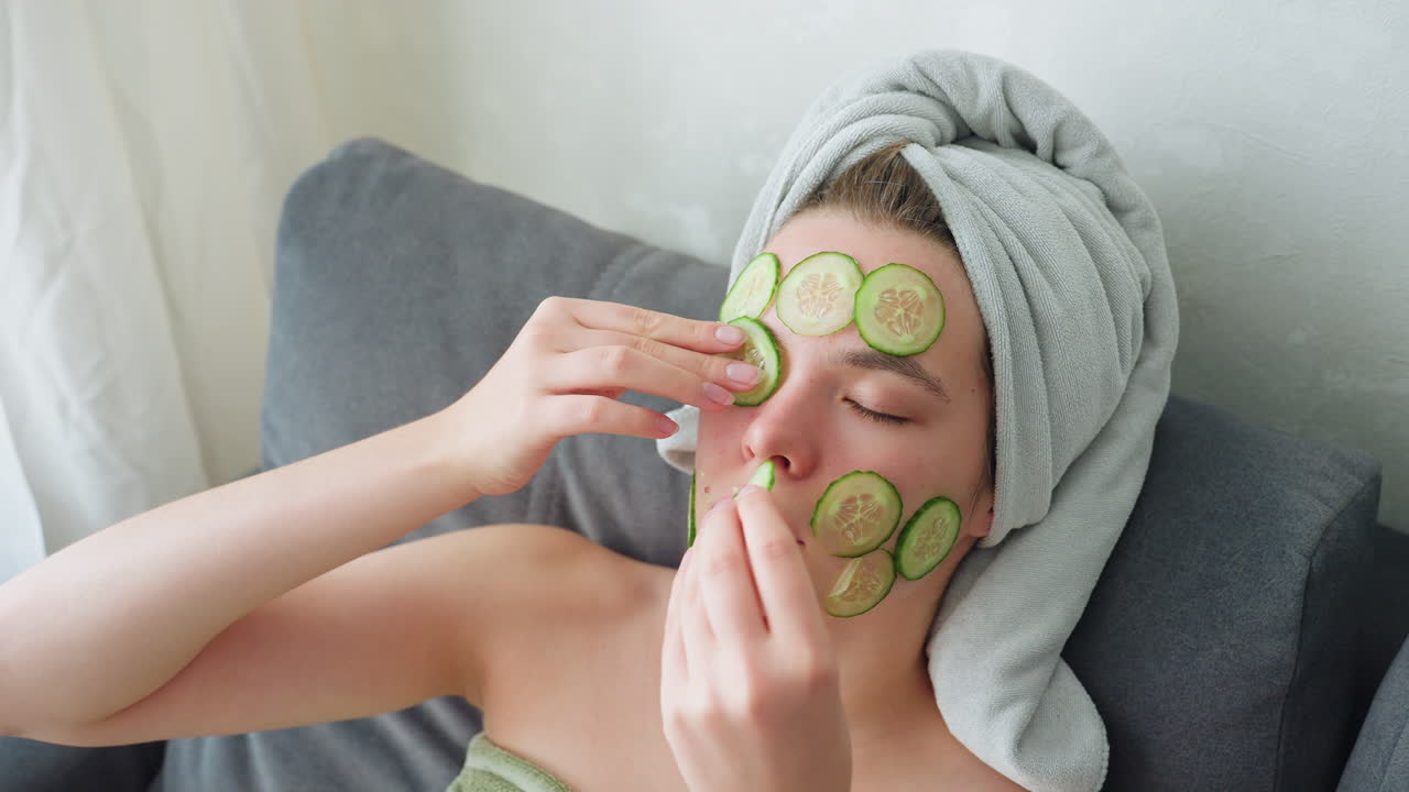 Close-up view of woman with towel wrapped around head placing cucumber slices over eyes and face in bright indoor setting, enjoying peaceful skincare and self-care moment focused on hydration