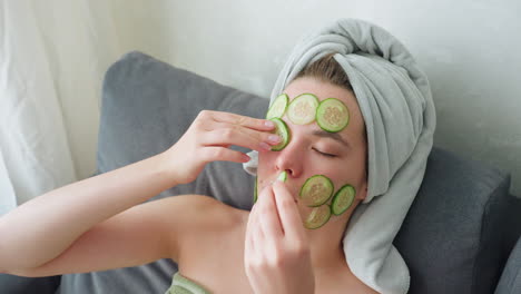 close-up view of woman with towel wrapped around head placing cucumber slices over eyes and face in bright indoor setting, enjoying peaceful skincare and self-care moment focused on hydration