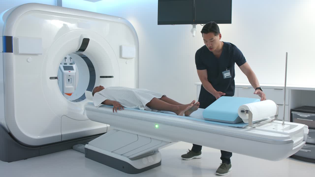 Paziente donna sdraiata sul lettino dello scanner, un tecnico asiatico posiziona un cuneo blu sotto i polpacci, preparando la scansione.