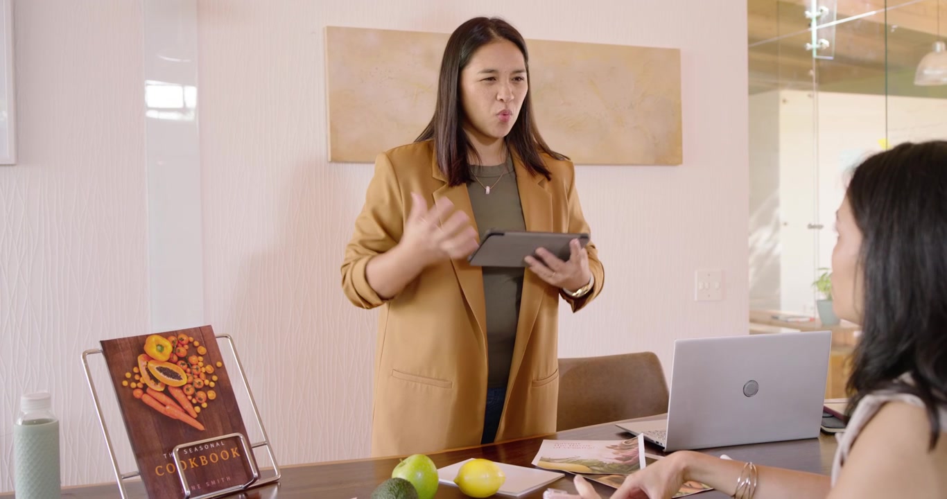 Asian female coworkers meeting at work table mature lead tapping tablet pointing to cookbook review