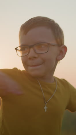 a young boy wearing a yellow shirt smiles shyly while moving his hands in a playful manner, standing in a sunlit grassy field during a serene sunset, capturing the essence of a joyful, carefree moment