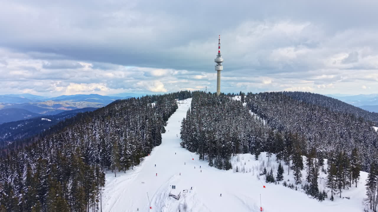 Cena de montanha nevada com esquiadores perto de uma torre alta em uma paisagem de inverno, cercada por árvores e montanhas durante o dia.