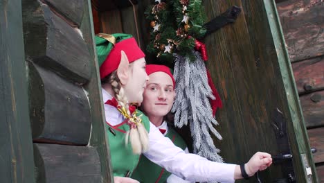 2 elves in green hats peep out of the door of the santa claus residence's wooden house in surprise