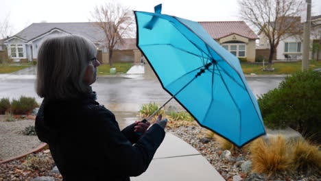 an old woman opening a blue umbrella and walking out into a winter storm as rain drops fall in slow motion