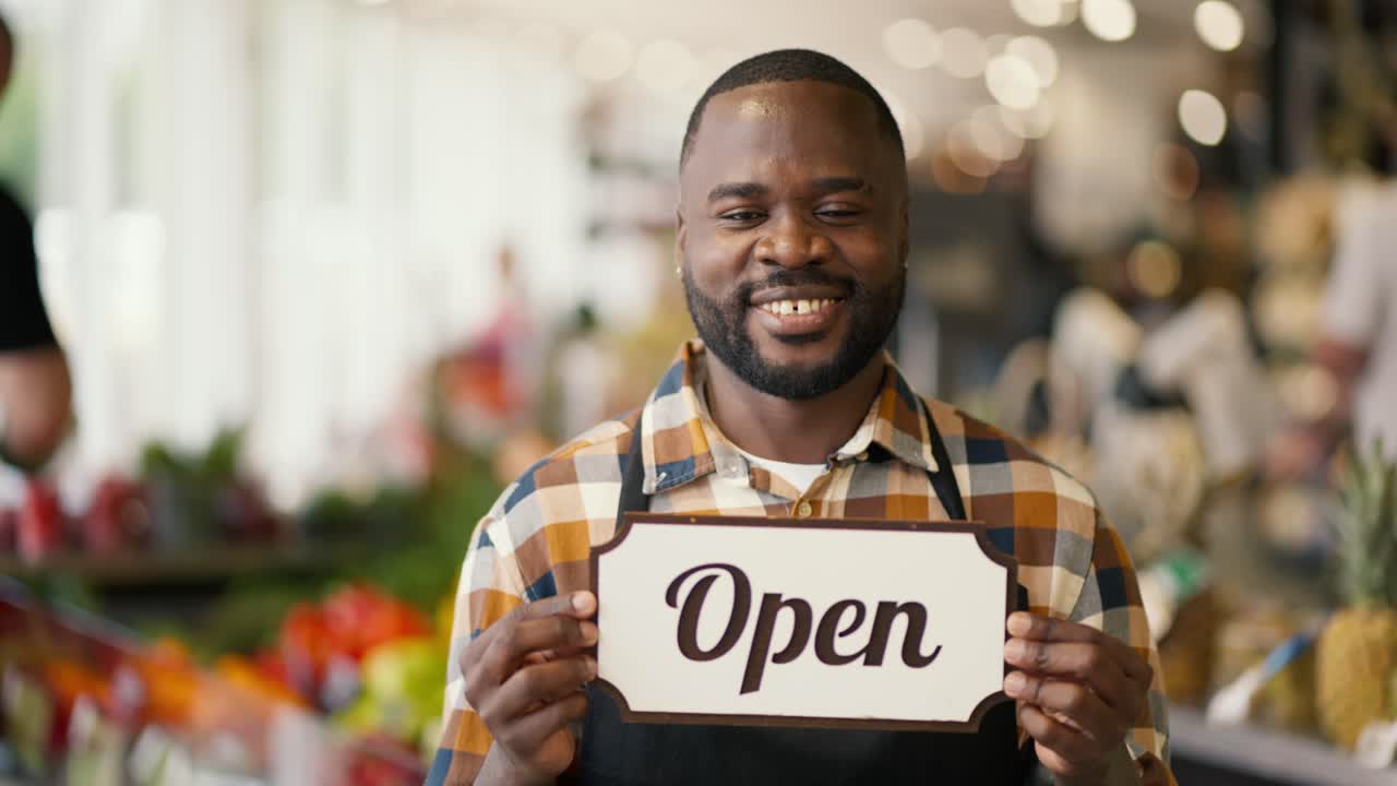 Portrait of a Black-skinned man in a plaid shirt and black apron holding a sign with the sign Open in a supermarket
