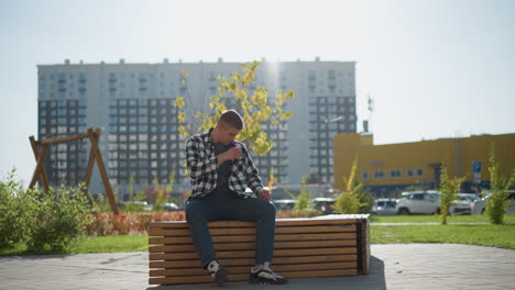 young man sits alone on wooden bench outdoors in urban park environment bends forward preparing to take pill with bright sunlight in background swaying tree leaves and blurred cars in distance