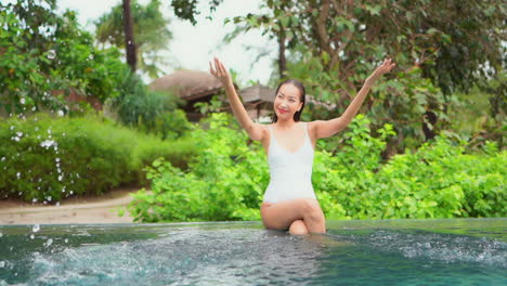 fit thai woman sitting on the edge of the pool and splashing swimming pool water with her hands towards the camera at an exotic tropical resort in a greenery background