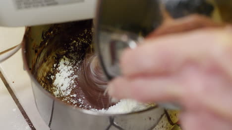 un chef en una cocina que usa una batidora eléctrica para hacer glaseado de chocolate para un pastel en un tazón de mezcla de metal con azúcar de confitería