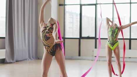 dos chicas jóvenes en leotardo practicando gimnasia rítmica con una cinta durante un ensayo en un estudio