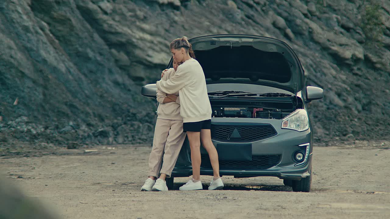 Two women comforting each other next to a broken down car