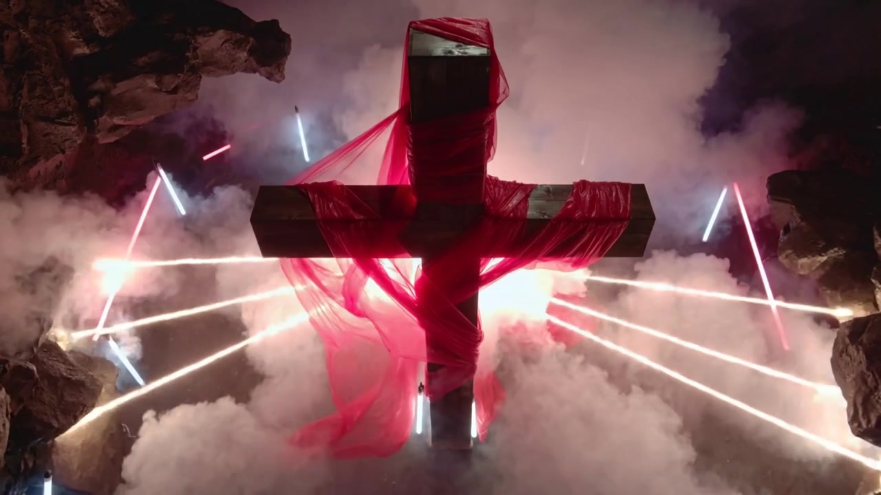 Des étincelles s'allument, envoyant des faisceaux radiaux derrière une grande croix en bois drapée de tissu rouge dans une grotte pour l'effet.