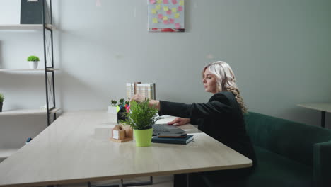 woman on green couch brings laptop forward and opens it to begin work in modern office, desk features potted plant, notebooks, phone, model visible, bright minimalist decor
